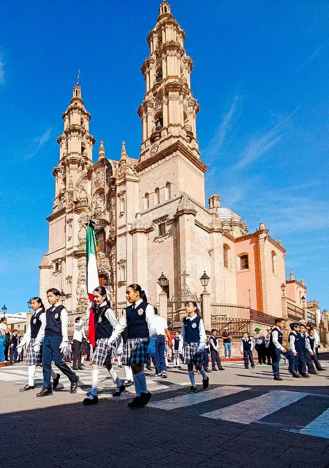 Estudiantes uniformados del colegio llevan la bandera mexicana durante un desfile frente a un templo de dos torres.