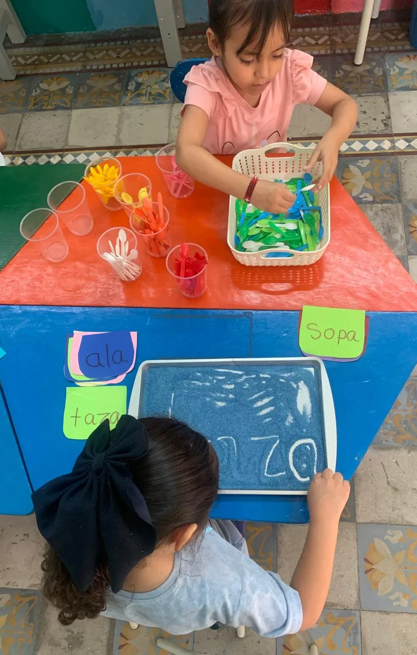 Dos alumnas clasifican cucharas de colores y realizan trazos en una bandeja con material fino.