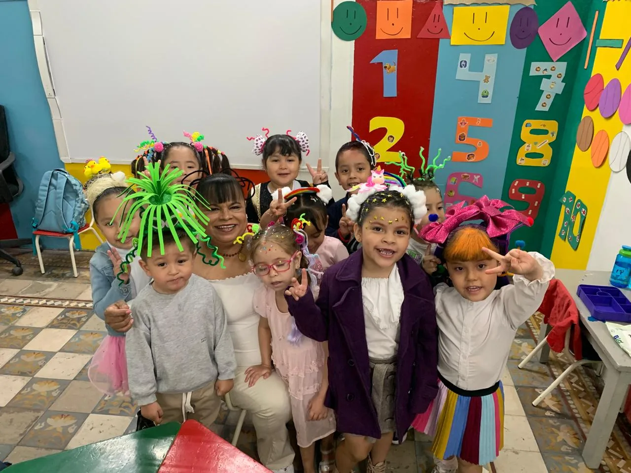 Niñas y niños con peinados creativos posan de frente junto a una acompañante en un aula de colores.
