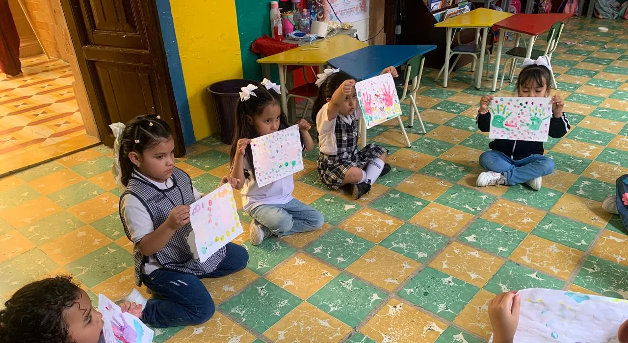 Alumnado con uniforme muestra sus trabajos de pintura en una actividad de aula.